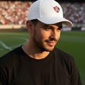 A man wearing a white Atlas FC Twill baseball cap with a red and black shield logo, standing on a green soccer field with a stadium in the background.