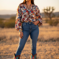 A woman standing in a dry grass field at sunset wearing a cream cowboy hat, a long-sleeve button-up blouse with a red, blue, and yellow floral print, blue jeans, and colorful embroidered cowboy boots.