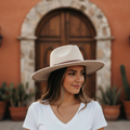 A woman wearing a wide-brimmed cream fedora hat and a white V-neck t-shirt, standing in front of a rustic terracotta wall and wooden door.