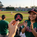 A person at a baseball stadium holds up a round black patch featuring a red letter A, with a smiling woman in a green Los Angeles Angels jersey and cap clapping in the background.