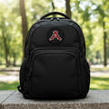 A black backpack with a circular patch featuring a red letter 'A' on a white background, sitting on a stone ledge with a blurred green park background.