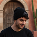 A man wearing a black knit beanie with a small white embroidered Aguascalientes logo and a black t-shirt, standing outdoors in front of a warm-toned stone archway and cactus.
