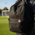 A person viewed from behind wearing a black backpack with a small red and Atlas FC white shield logo on the front pocket, standing on a green soccer field under a blue sky.