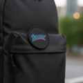 A close-up of a black backpack with a front utility pocket and a circular black patch featuring the brand name 'Bravos' in stylized blue and pink lettering, set against a blurred outdoor city background.
