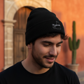 A man wearing a black knit beanie with a white embroidered Campeche logo, photographed outdoors in front of a warm-toned building with a cactus in the background.