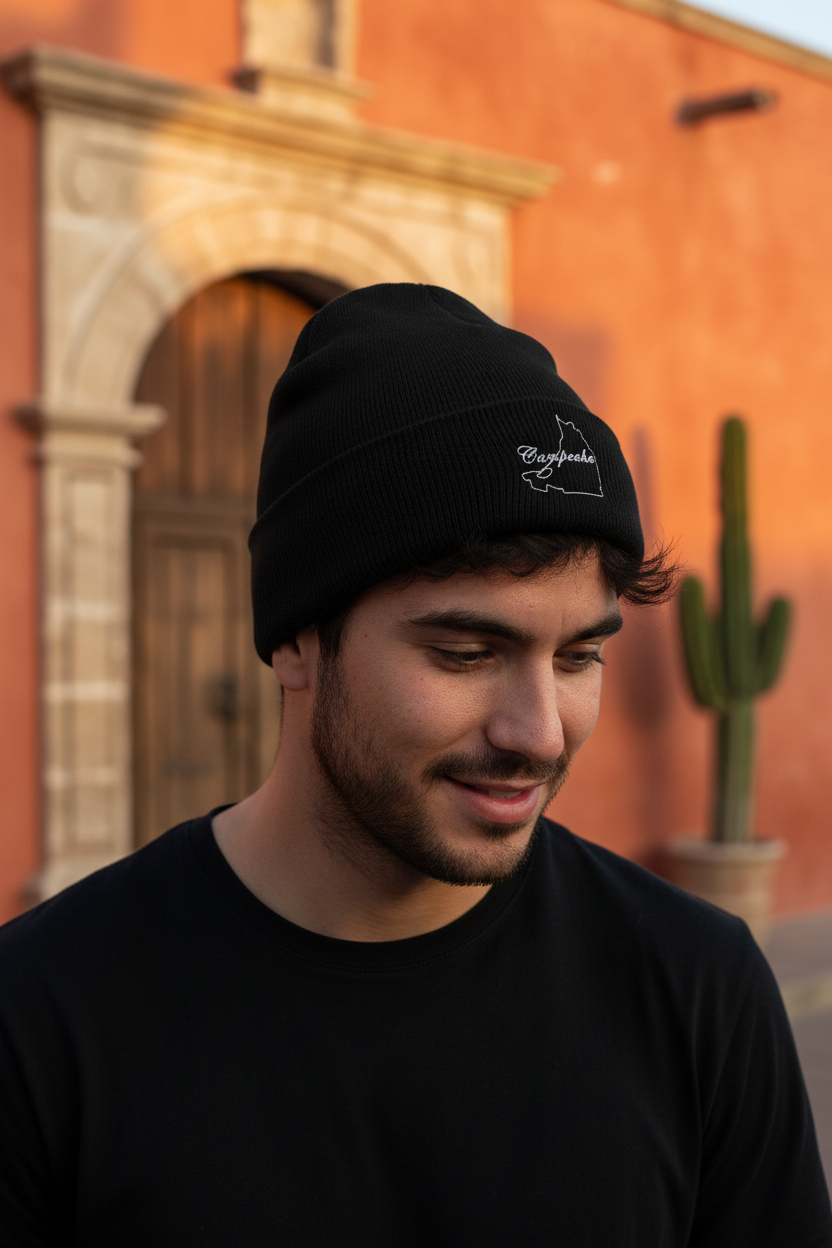 A man wearing a black knit beanie with a white embroidered Campeche logo, photographed outdoors in front of a warm-toned building with a cactus in the background.