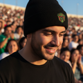 A man wearing a black knit beanie with a green and Ciudad Juarez crest logo on the front, photographed outdoors in front of a blurred crowd under a blue sky.