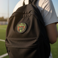 A person wearing a white t-shirt and blue jeans is shown from behind carrying a black backpack on a sports field. The backpack features a circular embroidered patch with the text 'JUAZ' and a green and red crest.