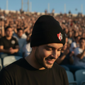 A man wearing a black knit beanie with a red and white Atlas logo, photographed in a stadium crowd at sunset.