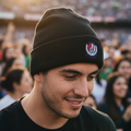 A man wearing a black knit beanie with a Club Atletico de San Luis logo, photographed in a stadium crowd during golden hour.