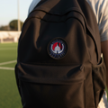 A black backpack worn by a person on a sports field, featuring a circular Club Atletico de San Luis crest patch on the front.