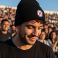 A man wearing a black knit beanie with a circular Club Cruz Azul emblem patch on the cuff, photographed outdoors in front of a crowd under a clear blue sky.
