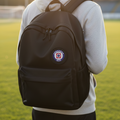 A person viewed from behind wearing a black backpack with a circular red, white, and blue Cruz Azul crest patch on the back, standing on a grassy field at sunset.
