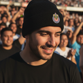 A man in a black beanie with a colorful Club Guadalajara Chivas crest and a black t-shirt, photographed in a stadium crowd at sunset.