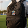 A black backpack with a circular Chivas soccer crest patch on the front, worn by a person standing on a green soccer field at sunset.