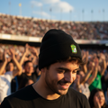 A man in a black Club Leon Cuffed beanie and black t-shirt smiling in a crowded outdoor stadium under a blue sky.
