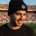 A man wearing a black knit beanie with a Club Mazatlan FC crest on the front, photographed outdoors at a stadium with blurred stands in the background.