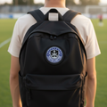 A person viewed from behind wearing a black backpack on a green sports field at sunset. The backpack has a circular blue and white Mazatlan FC crest patch on the front.