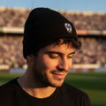 A man wearing a Club Monterrey black knit beanie with a small shield logo on the front, standing on a stadium field at sunset.