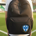 A person viewed from behind wearing a black backpack on a green soccer field. The backpack has a large front zip pocket with a circular blue and white embroidered Club Monterrey logo patch featuring yellow stars.