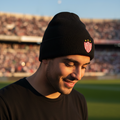A man wearing a black knit beanie with the Club Necaxa crest, standing on a soccer field with a stadium in the background.