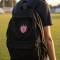 A person wearing a black backpack with a red and white FC Necaxa crest patch on the front, standing on a grassy field at sunset.