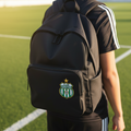 A black backpack with a green and white FC Pachuca crest patch on the front pocket, worn by a person standing on a green sports field.