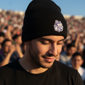 A man wearing a black knit beanie with a white and purple embroidered Club Puebla crest, photographed outdoors with a blurred crowd in the background.