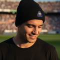 A man wearing a black knit beanie with a small blue and yellow Pumas UNAM logo, standing on a soccer field with a stadium in the background.