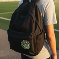 A person viewed from behind wearing a black backpack with a circular Club Queretaro crest patch on the front pocket, standing outdoors near a sports field at sunset.