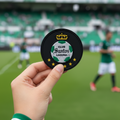 A hand holds a round embroidered Club Santos Laguna patch in the foreground, with a blurred soccer stadium and player in the background.