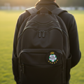 A person viewed from behind wearing a black backpack outdoors on a grassy field at sunset. The backpack has a front zip pocket with a round crest patch that reads 'Club Santos'.