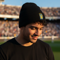 A man wearing a black knit beanie with a small Club Tigres yellow and blue crest patch on the front, standing in a stadium with blurred spectators in the background.