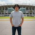 A man wearing a light gray t-shirt, dark blue jeans, a black Club Tijuana bucket hat, and white sneakers stands in front of a large stadium under a cloudy sky.