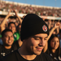 A man in a Club Tijuana black beanie with a circular red, white, and black logo stands in a crowded outdoor stadium at sunset, wearing a black t-shirt. The background shows a blurred crowd and stands under a blue sky.