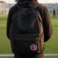 A person viewed from behind wearing a black hoodie and black backpack on a grassy sports field. The backpack has a circular red, white, and black crest Club Tijuana patch on the lower front pocket.
