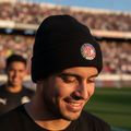 A man wearing a black knit beanie with the Club Toluca crest, standing on a soccer field with stadium stands blurred in the background.