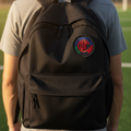 A person viewed from behind wearing a black backpack with a circular red, green, and blue crest Club Toluca FC patch on the upper back, standing outdoors on a grassy field at sunset.