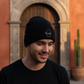A man wearing a black knit beanie with a small white embroidered Coahuila logo and a black t-shirt, standing outdoors in front of a warm-toned stone archway and cactus.