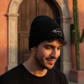 A man wearing a black knit beanie with a white embroidered Colima logo and a black t-shirt, standing outdoors in front of a warm-toned stucco building with a wooden door and a cactus.