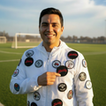 Man standing on a soccer field wearing a white jacket covered in embroidered patches, pointing at a black patch with red “Diablos” text.