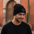 A man wearing a black knit beanie with a white embroidered Durango logo and a black t-shirt, standing outdoors in front of a warm terracotta-colored building with a wooden door and a cactus.