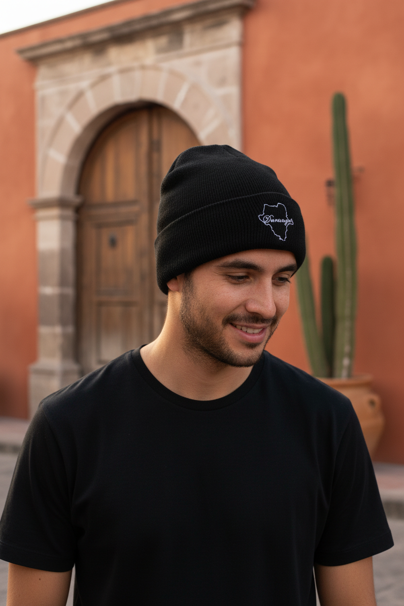 A man wearing a black knit beanie with a white embroidered Durango logo and a black t-shirt, standing outdoors in front of a warm terracotta-colored building with a wooden door and a cactus.