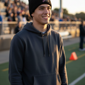 A smiling young man standing on a sports field at sunset, wearing El Aguila de Veracruz a black knit beanie with a small red and white logo and a dark charcoal hoodie.
