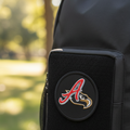 A close-up of a black backpack with a front pocket featuring a circular patch with a red and gold Atlanta Braves 'A' logo, set against a blurred outdoor park background.