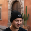 A man wearing a black knit beanie with a white Guanajuato embroidered logo, standing outdoors in front of a warm-toned stone building.