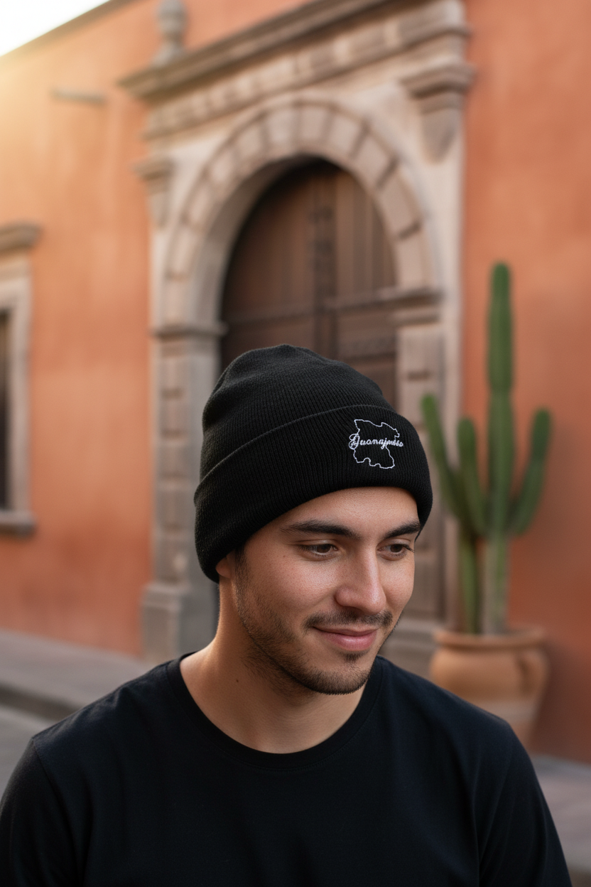 A man wearing a black knit beanie with a white Guanajuato embroidered logo, standing outdoors in front of a warm-toned stone building.