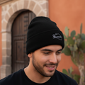 A man wearing a black knit beanie with a small white Guerrero embroidered logo, photographed outdoors in front of a terracotta-colored building with a wooden door and cactus. 