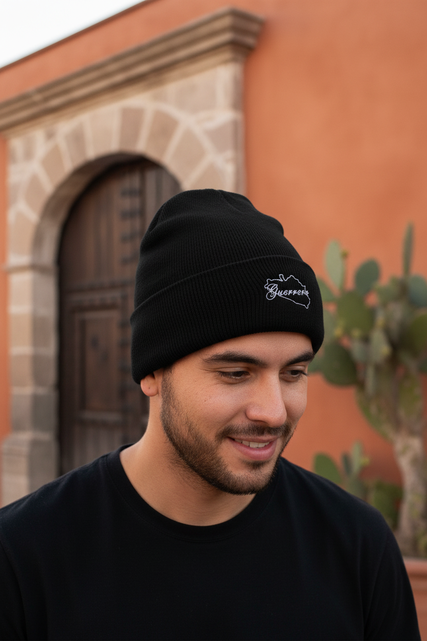 A man wearing a black knit beanie with a small white Guerrero embroidered logo, photographed outdoors in front of a terracotta-colored building with a wooden door and cactus. 