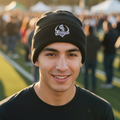 A smiling young man outdoors wearing a black knit beanie with a white and gray Guerreros de Oaxaca logo and a black t-shirt, with a blurred crowd and event tents in the background.